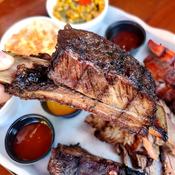 A hand holds up a large, glistening Santa Maria BBQ beef rib, with sides and sauces blurred in the background.